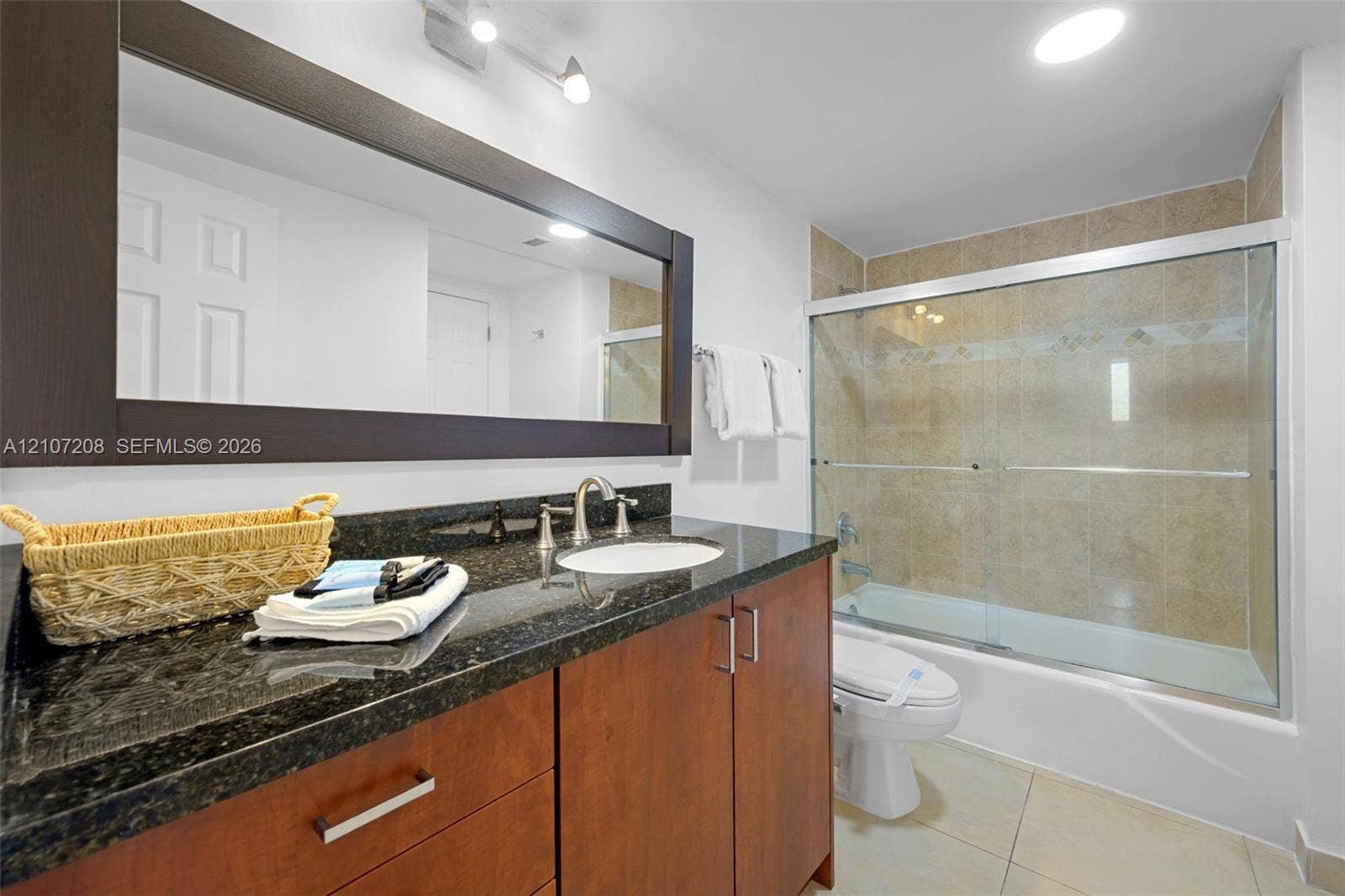 19370 Collins Avenue, #1405, Sunny Isles Beach, Sunny Isles Beach, FL 33160 Bathroom featuring a wood-finish vanity with a dark stone countertop, a large framed wall mirror, and a tub-shower combination with tiled surround and glass sliding doors