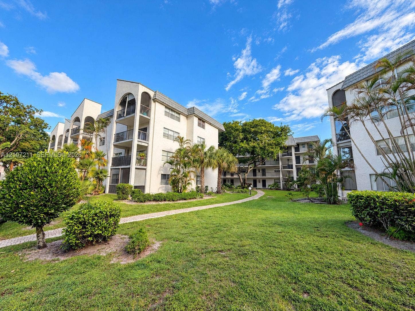 Multi-story exterior featuring arched architectural detailing, private balconies with metal railings, a paved pedestrian pathway, and mature landscaping