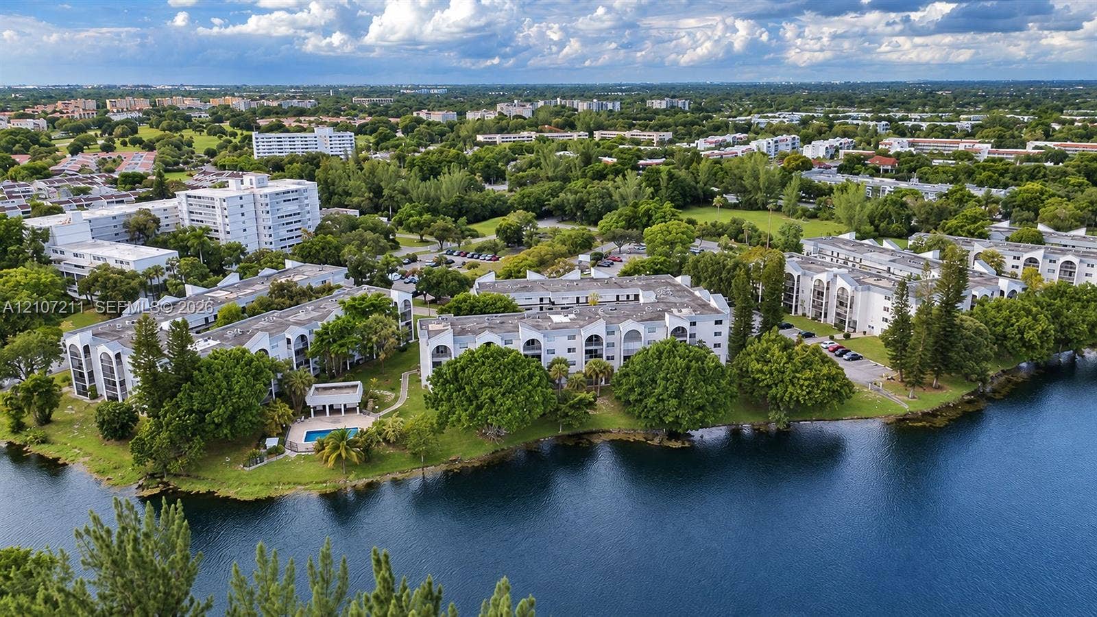 Aerial perspective of multi-story residential buildings featuring white stucco facades, arched balcony detailing, a waterfront location, mature trees, and an on-site swimming pool