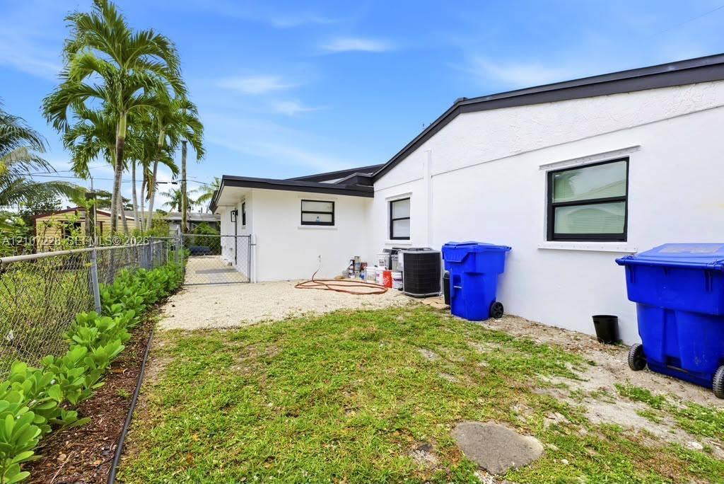 Exterior side yard featuring white stucco siding, dark-trimmed windows, a chain-link fence, and mature palm trees