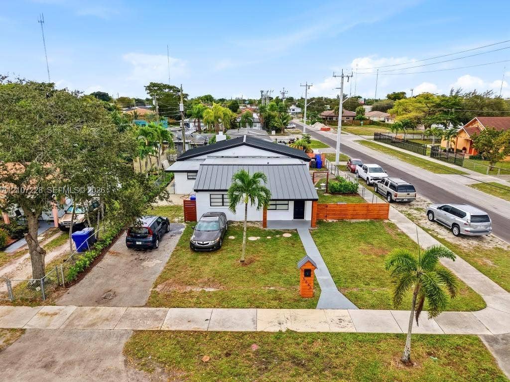 Single-story exterior featuring a metal roof, front yard with mature palm trees, concrete driveway, and wood-slat perimeter fencing