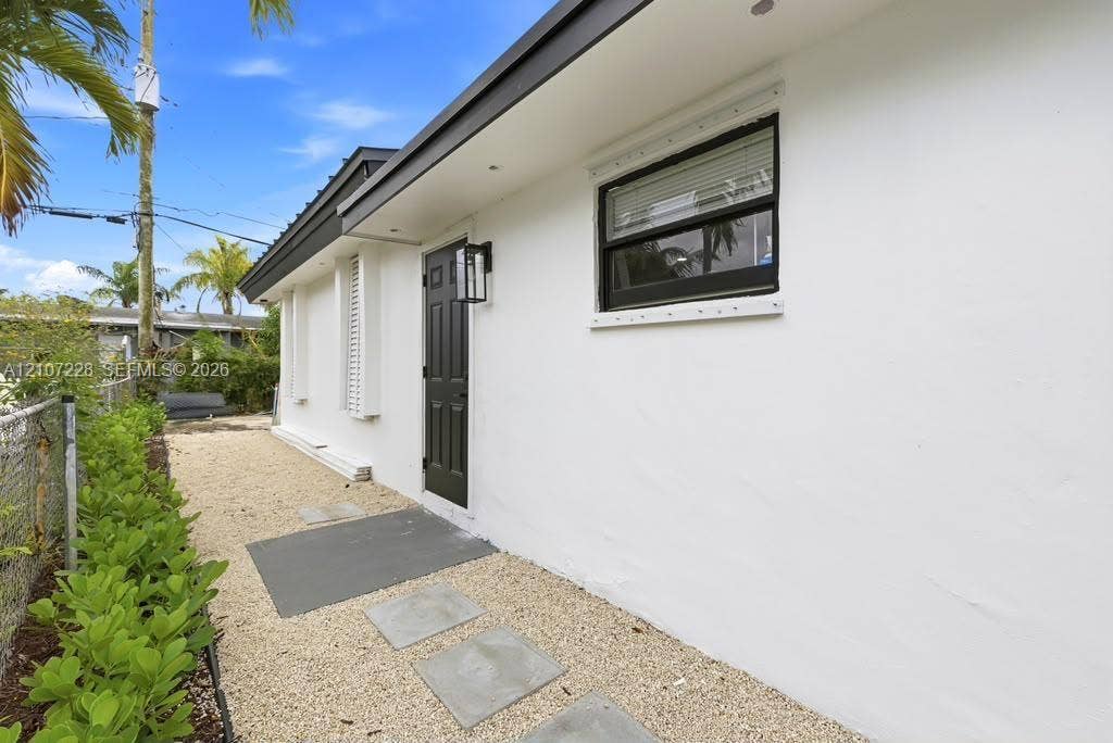 Exterior side entrance featuring a dark-toned door with a modern rectangular wall sconce, white smooth-finish siding, a gravel walkway with concrete paver accents, and a low-profile flat roofline