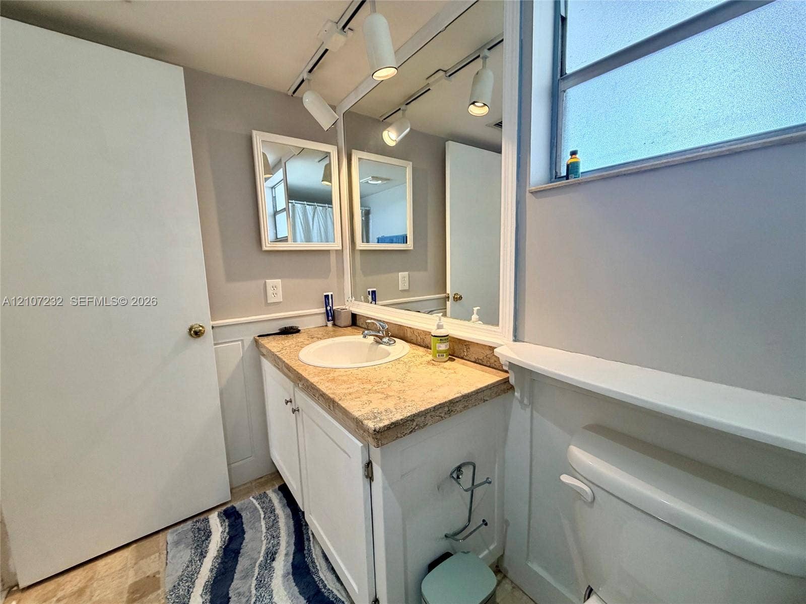 Vanity featuring a stone-look countertop with an integrated basin, white shaker-style cabinetry, wall-to-wall mirror, track lighting, and a window ledge