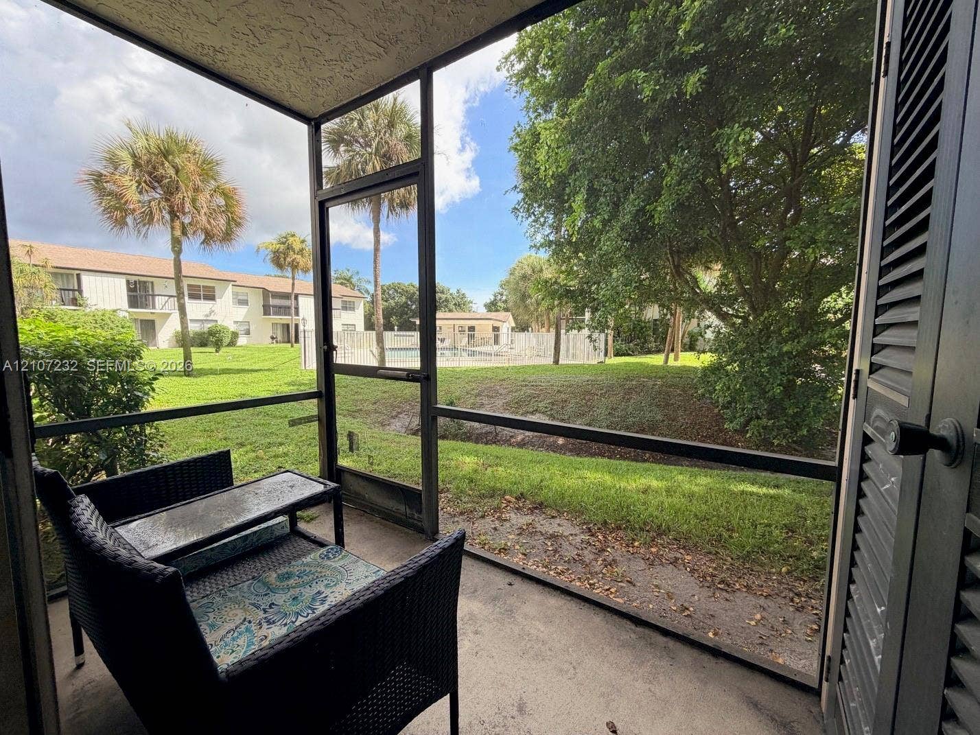 Screened-in patio with concrete flooring, louvered exterior door, and metal framing, overlooking landscaped grounds with mature palm trees