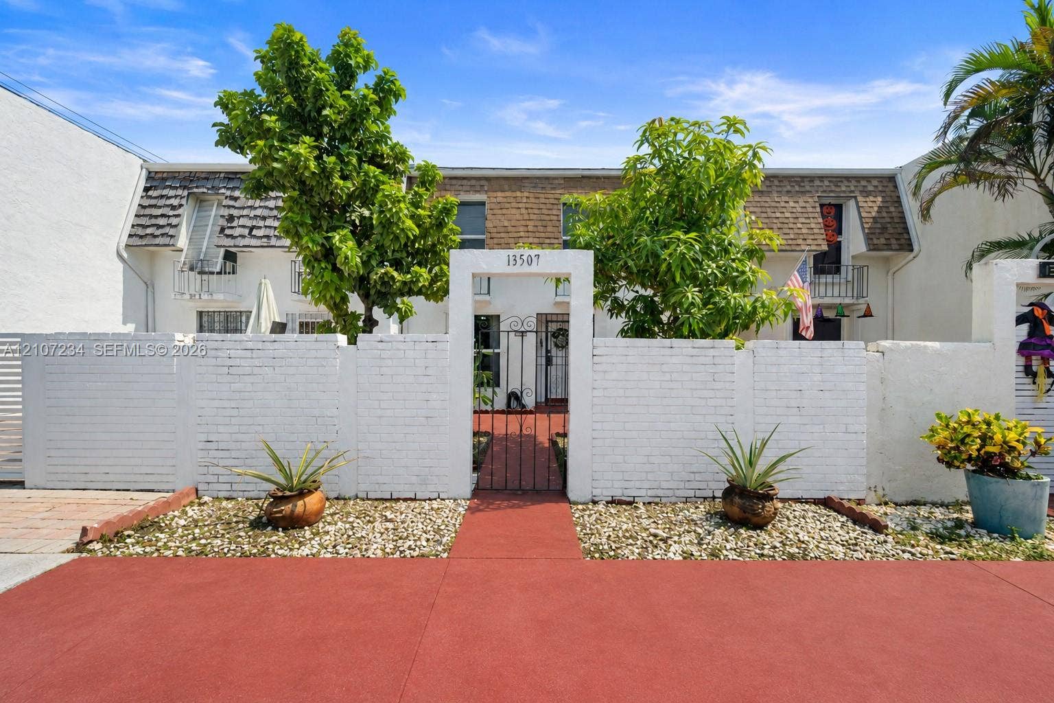 13507 NE 24th Court, North Miami, FL 33181 Two-story townhouse exterior featuring a white-painted brick privacy wall, a gated entryway, shingled mansard roof detailing, and individual second-story balconies
