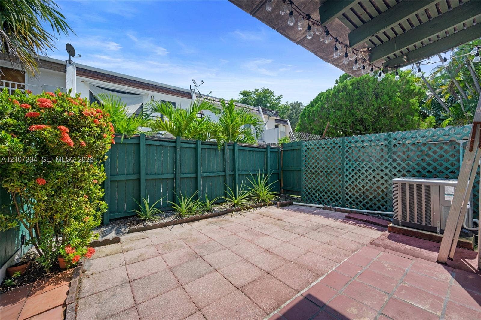 13507 NE 24th Court, North Miami, FL 33181 Private patio featuring terracotta-style paving, emerald-green wood privacy fencing, decorative lattice screening, and established tropical landscaping