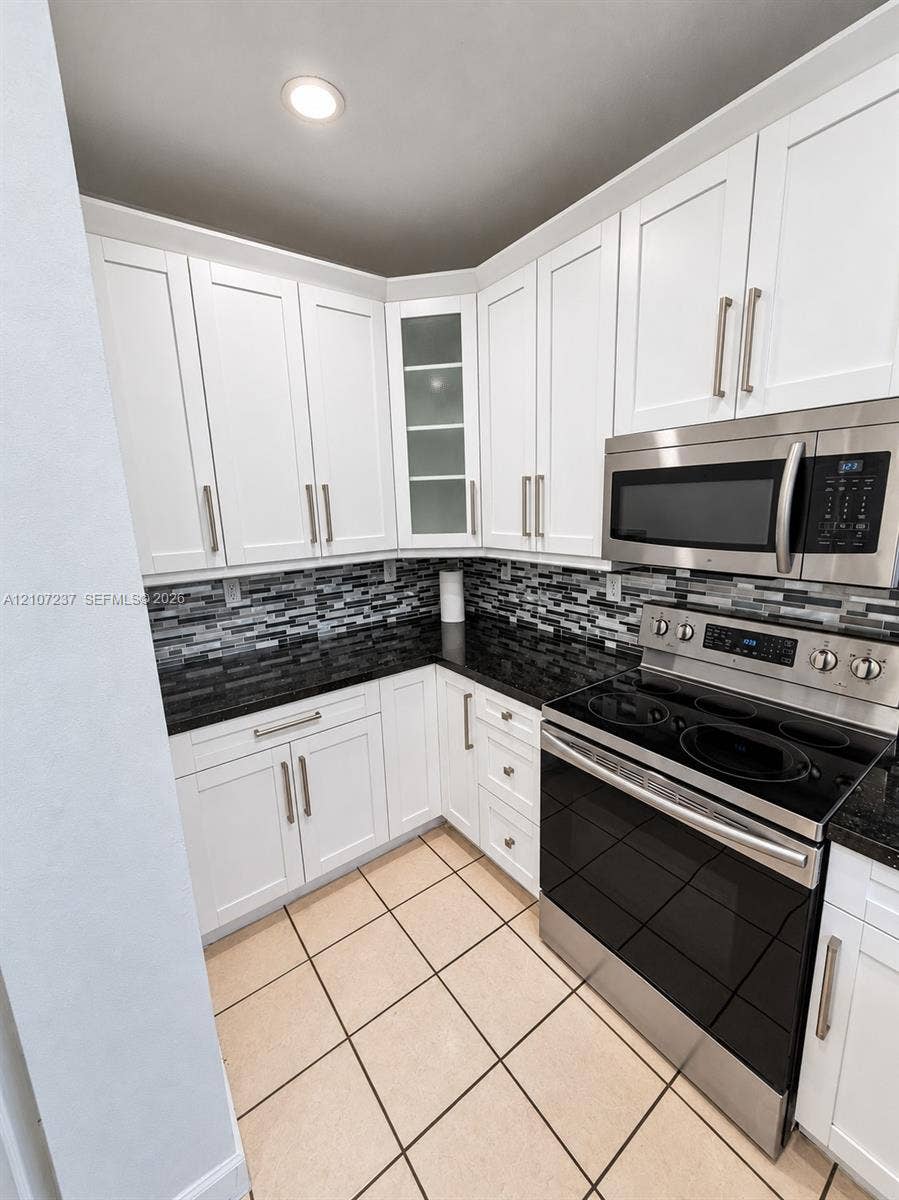 2529 NE 41st Terrace, Homestead, FL 33033 White shaker-style cabinetry with glass-front display accents, dark stone-finish countertops, mosaic tile backsplash, stainless steel appliances, and tile flooring