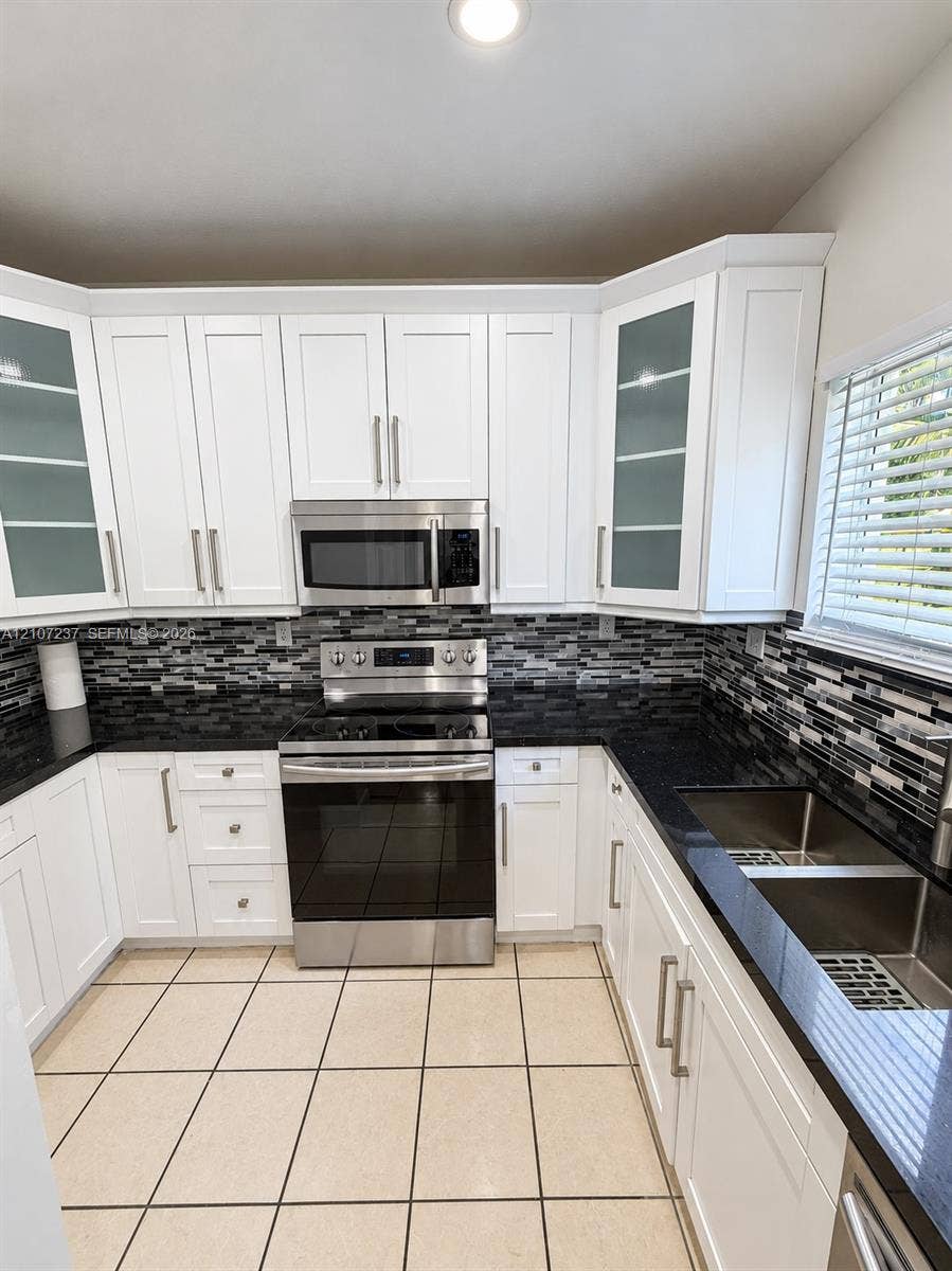 2529 NE 41st Terrace, Homestead, FL 33033 White shaker-style cabinetry with glass-front uppers, stainless steel appliances, dark stone countertops, mosaic tile backsplash, and dual-basin stainless steel sink