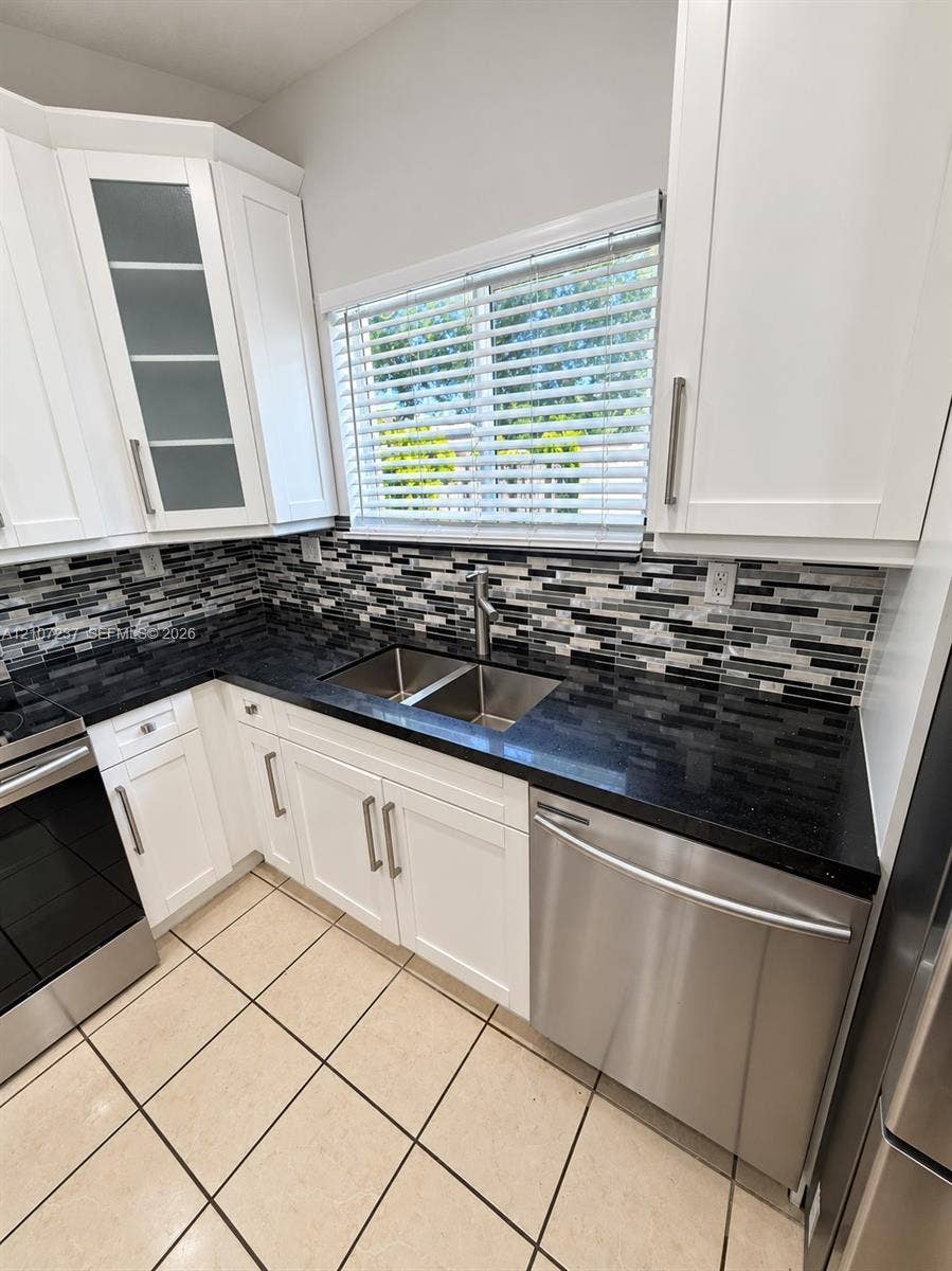 2529 NE 41st Terrace, Homestead, FL 33033 Kitchen featuring white shaker-style cabinetry with glass-front accents, dark stone countertops, a multi-tonal linear tile backsplash, stainless steel appliances, and neutral tile flooring