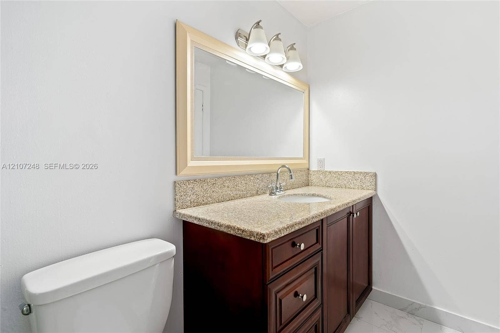 9983 W Atlantic Boulevard, #9983, Coral Springs, Coral Springs, FL 33071 Vanity featuring dark-stained wooden cabinetry, stone-finish countertop with an integrated basin, framed wall mirror, three-light fixture, and polished metal faucet