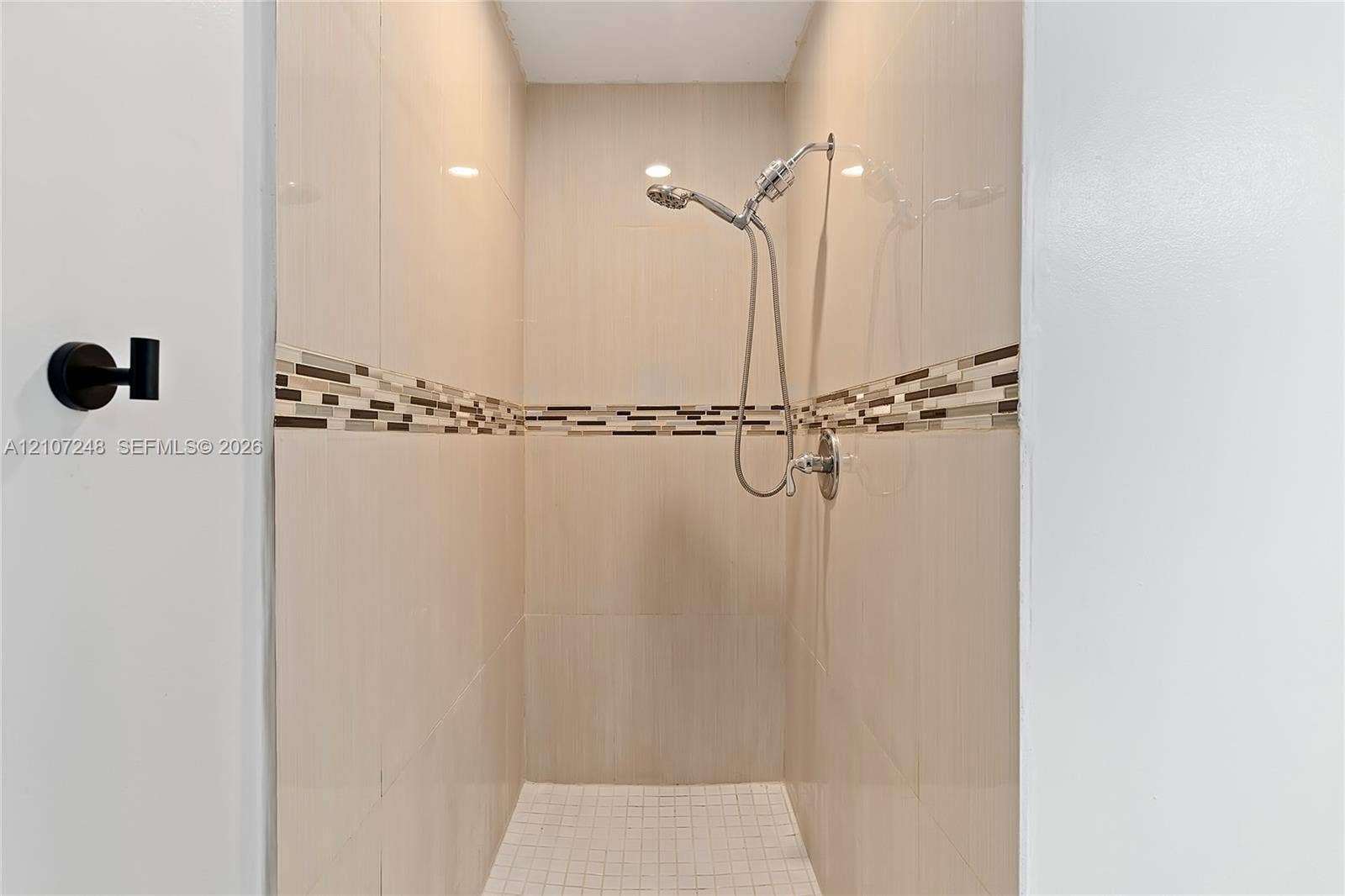 9983 W Atlantic Boulevard, #9983, Coral Springs, Coral Springs, FL 33071 Shower stall featuring floor-to-ceiling neutral wall tile, a horizontal decorative glass mosaic accent border, a small-format square tile floor, and a polished chrome handheld shower fixture
