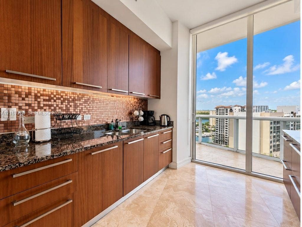 16001 Collins Avenue, #1605, Sunny Isles Beach, FL 33160 Kitchen featuring flat-panel wood-finish cabinetry, stone countertops, mosaic tile backsplash, floor-to-ceiling glass sliding doors, and large-format stone flooring