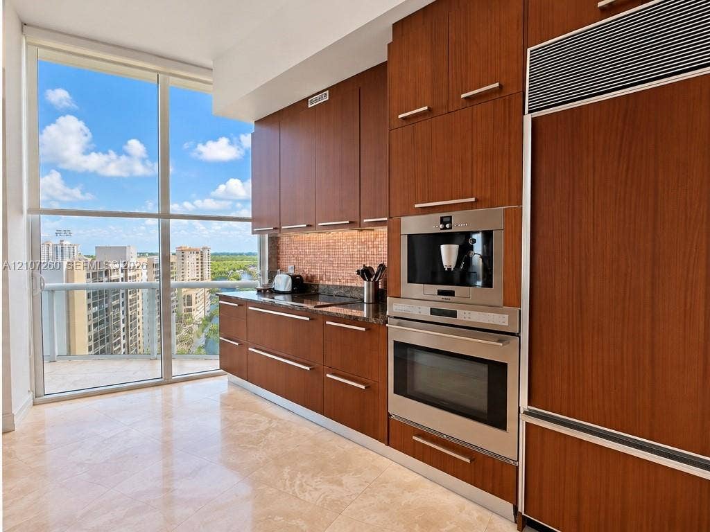16001 Collins Avenue, #1605, Sunny Isles Beach, FL 33160 Kitchen featuring custom flat-panel wood cabinetry, integrated stainless steel refrigerator, built-in wall oven and coffee station, stone countertops with a mosaic tile backsplash, and floor-to-ceiling windows