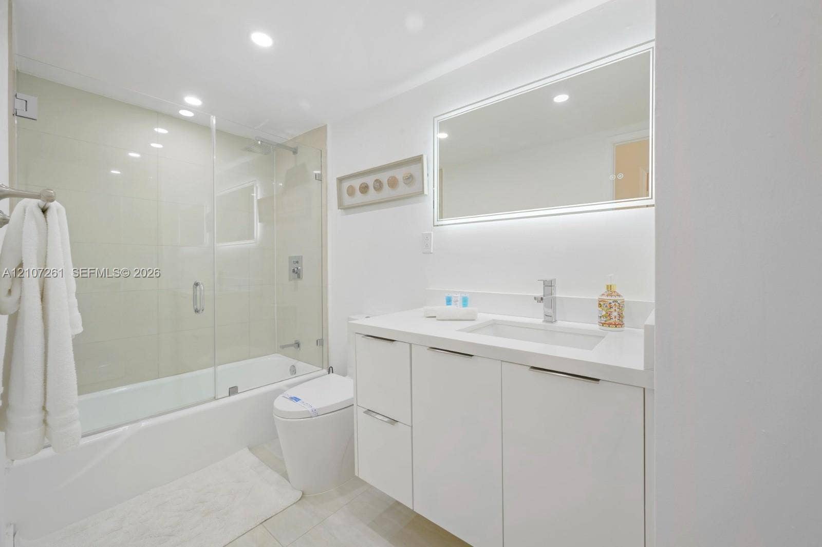 19370 Collins Avenue, #1601, Sunny Isles Beach, Sunny Isles Beach, FL 33160 Bathroom featuring a white floating vanity with a solid-surface countertop, an integrated rectangular sink, a glass-enclosed shower-tub combination, and an LED-backlit mirror