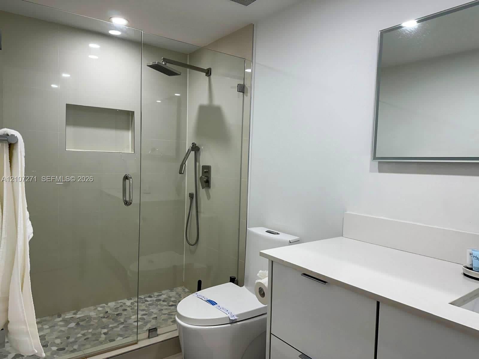Bathroom featuring a frameless glass walk-in shower with a recessed wall niche, pebble-stone flooring, rainfall showerhead, and a vanity with a solid-surface countertop