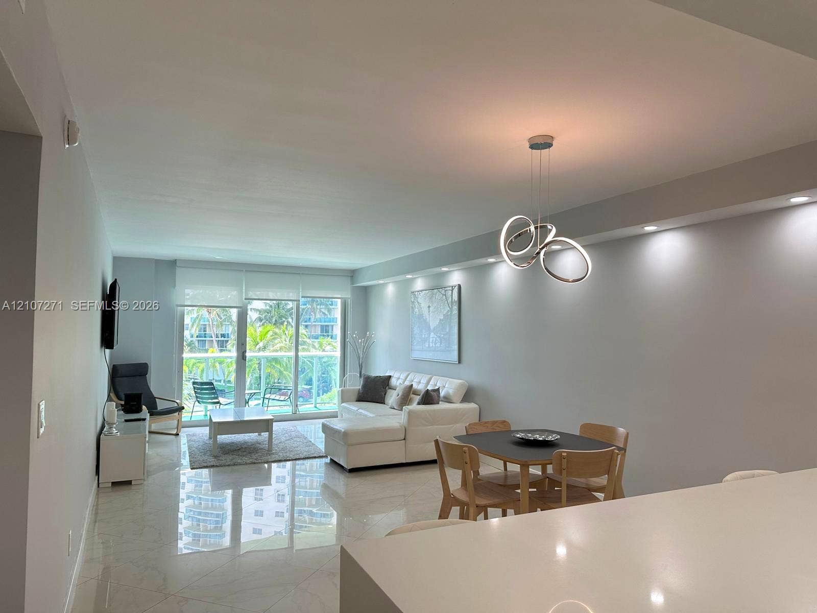 Open-concept area featuring polished tile flooring, recessed ceiling lighting, a modern sculptural chandelier, and a sliding glass door leading to a balcony with a glass railing