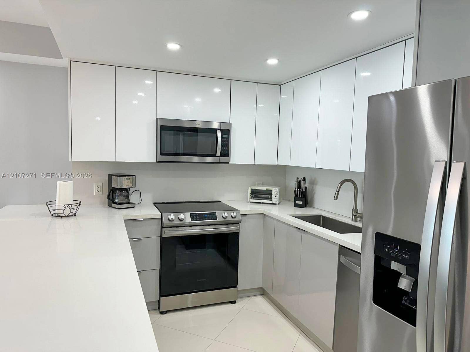 Modern kitchen featuring high-gloss white cabinetry, stainless steel appliances, a stone countertop, recessed ceiling lighting, and large-format tile flooring