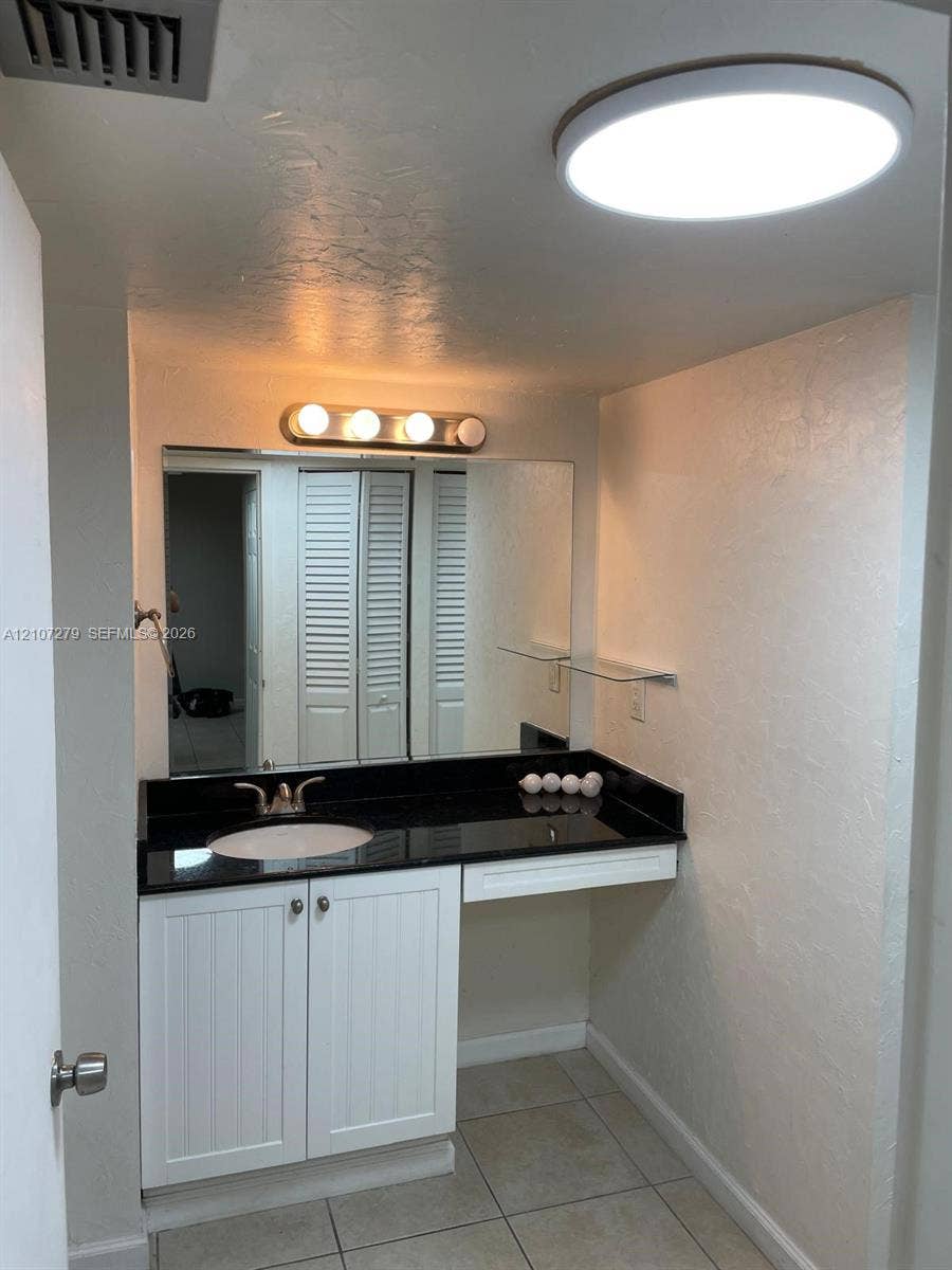 20100 W Country Club Drive, #601, Aventura, FL 33180 Vanity area featuring white shaker-style cabinetry, dark stone-finish countertop with an integrated basin, large rectangular mirror, and dual ceiling-mounted lighting fixtures