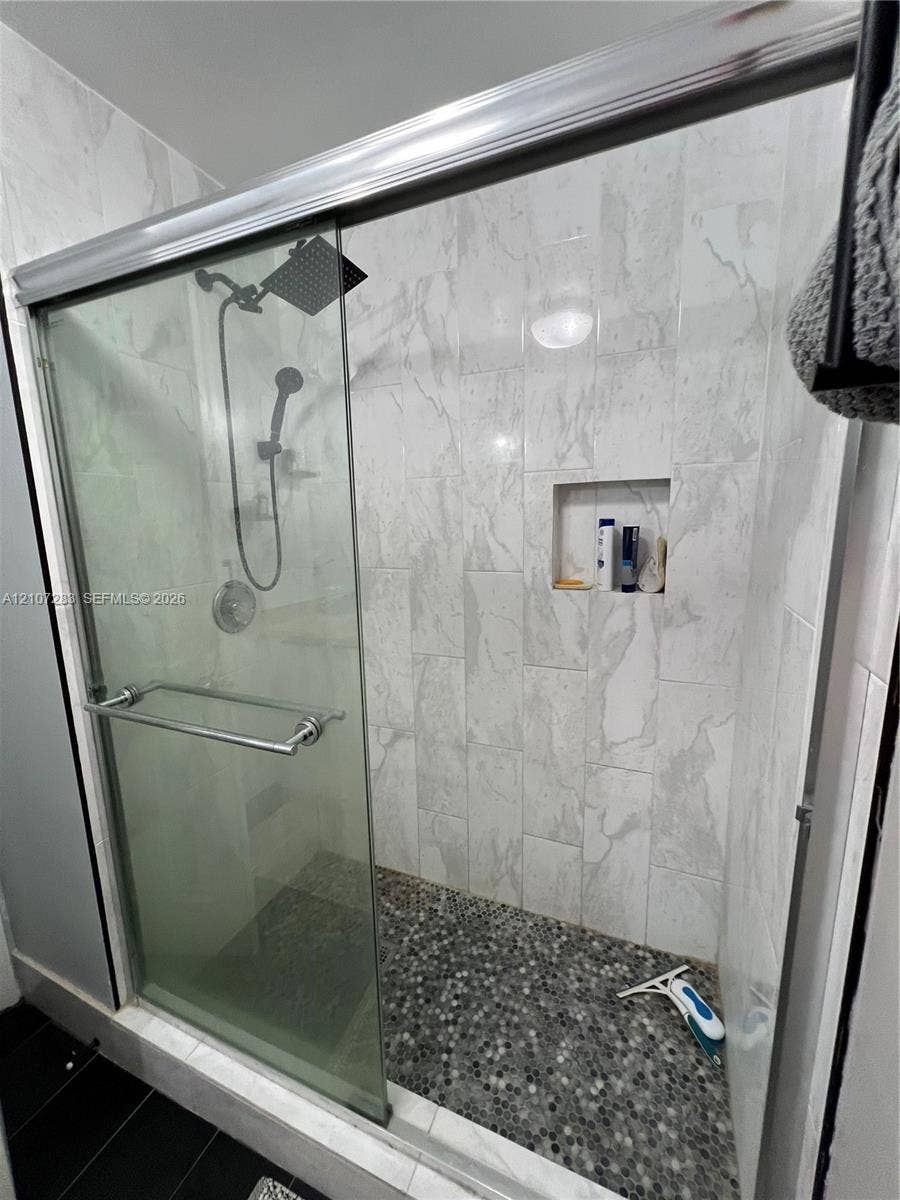 3301 Raleigh Street, #4E, Hollywood, FL 33021 Walk-in shower featuring glass sliding doors, large-format tile surround, integrated tiled niche, rainfall shower head with handheld attachment, and a pebble-stone floor