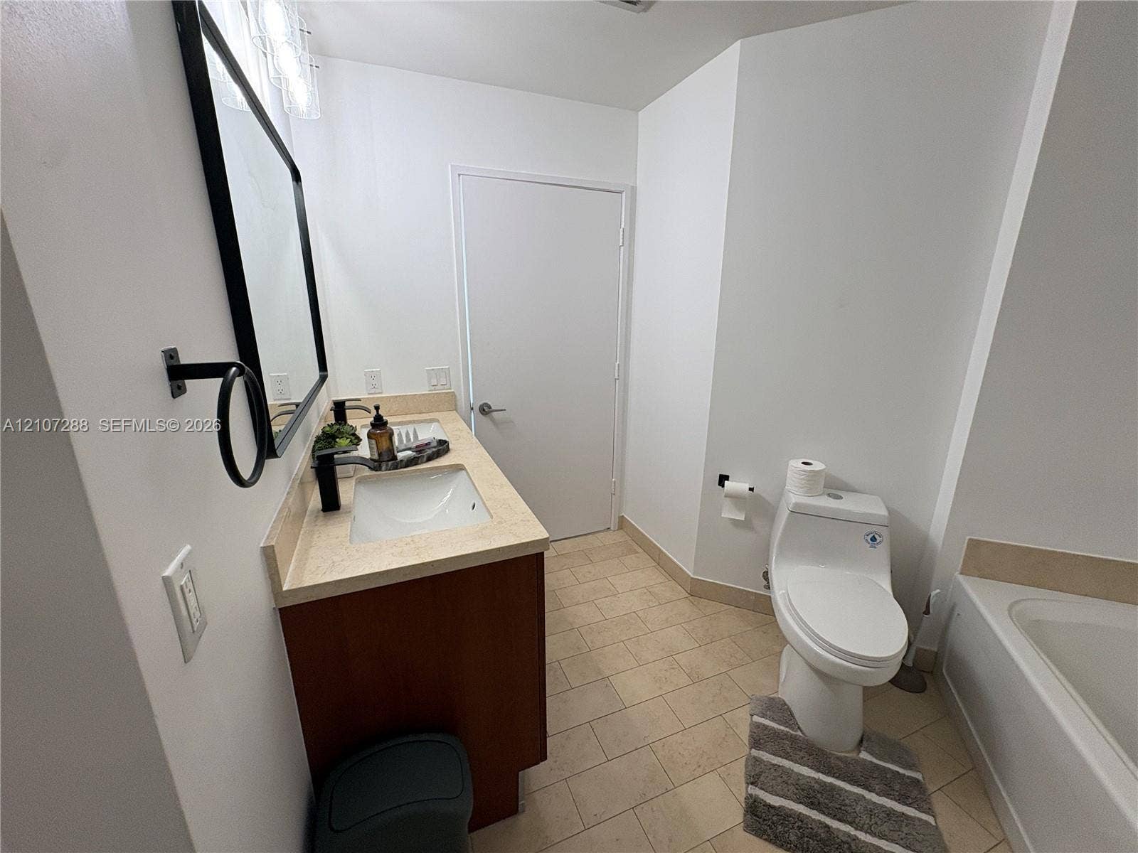 Bathroom featuring a wood-finish vanity with a stone-composite countertop, an integrated rectangular sink, a black-framed mirror, and ceramic tile flooring
