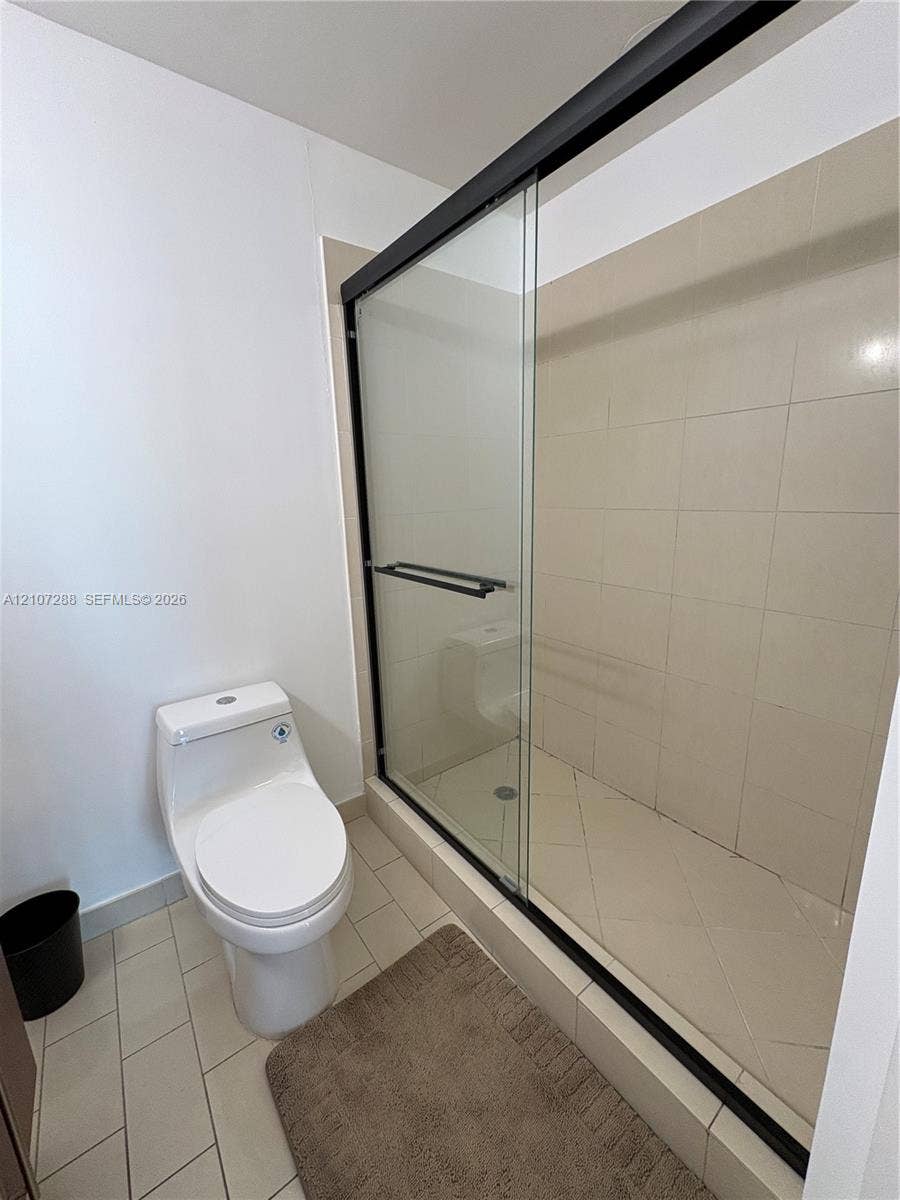 Walk-in shower featuring a sliding glass door with a black frame, ceramic tile surround, and neutral floor tile, paired with a white dual-flush toilet