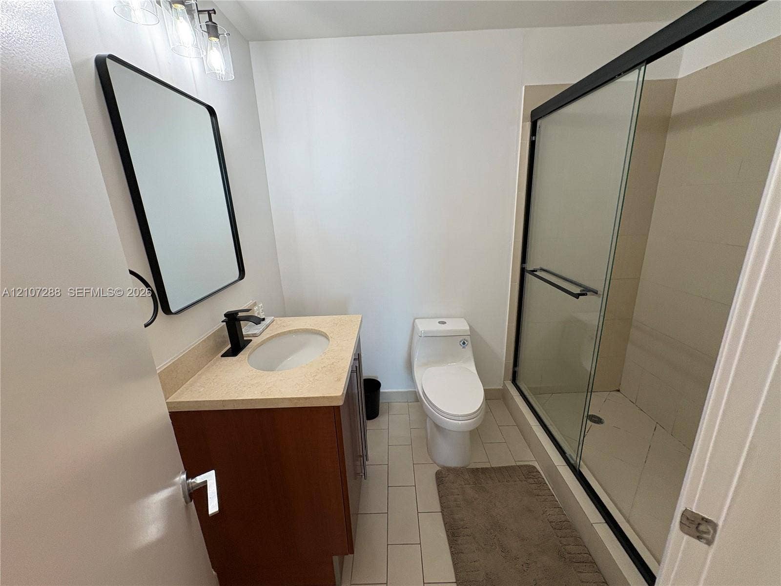 Wood-finish vanity with a light-toned countertop, undermount sink, matte black faucet, rectangular mirror with a black frame, and a walk-in shower featuring a sliding glass enclosure
