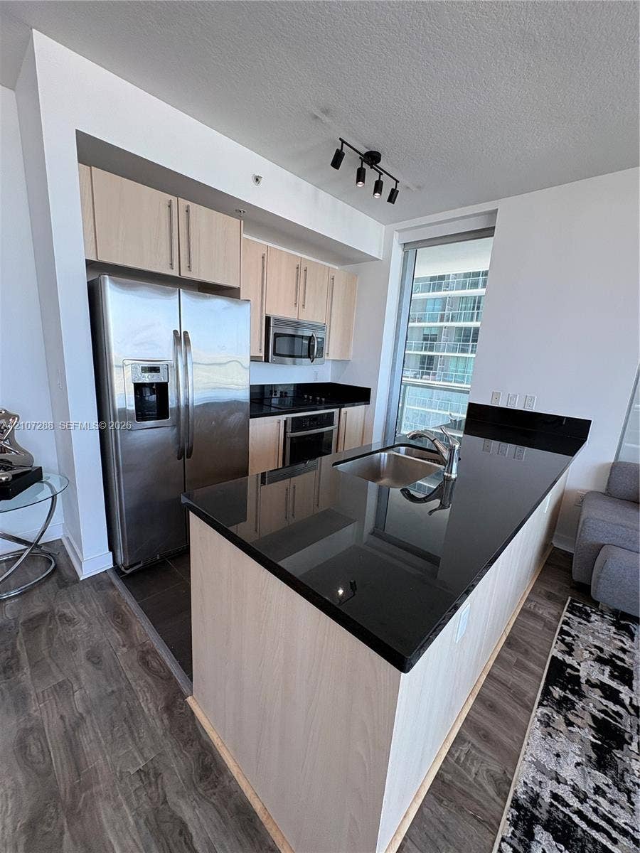 Kitchen featuring light wood-finish cabinetry, black stone countertops, stainless steel appliances, a center island with an undermount sink, and track lighting