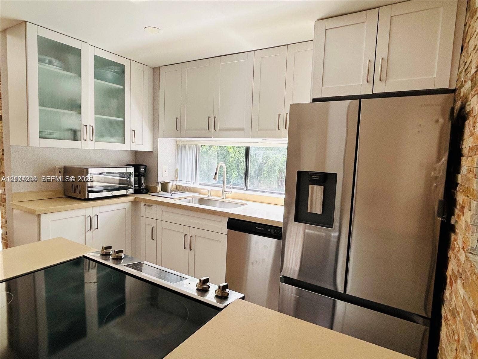 17021 N Bay Road, #219, Sunny Isles Beach, Sunny Isles Beach, FL 33160 Kitchen featuring white Shaker-style cabinetry with glass-front uppers, stainless steel appliances, a stone-textured accent wall, and a solid-surface countertop with an integrated cooktop