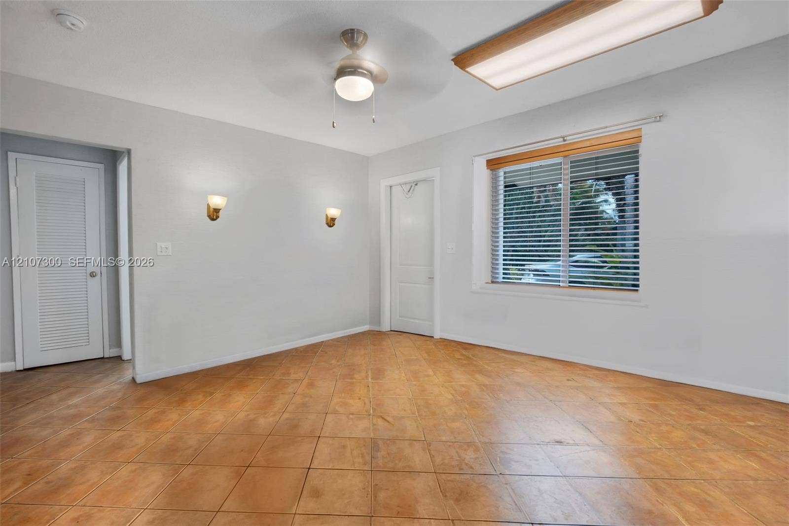 2051 NE 68th Street, Fort Lauderdale, FL 33308 Interior space featuring tile flooring, dual wall-mounted light fixtures, a ceiling fan with integrated light, a louvered door, and a recessed window with horizontal blinds