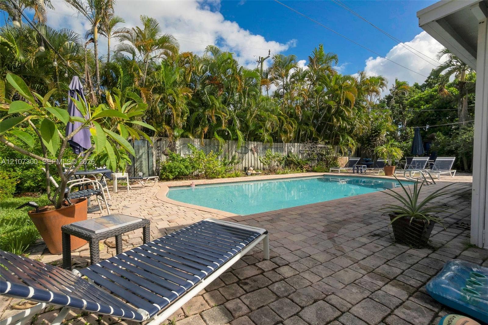 2051 NE 68th Street, Fort Lauderdale, FL 33308 Outdoor area featuring a rectangular in-ground swimming pool, expansive paver patio, and perimeter privacy fencing surrounded by mature palm trees