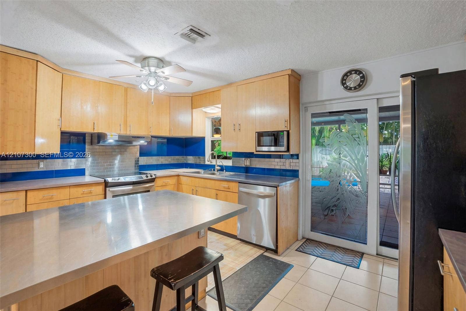 2051 NE 68th Street, Fort Lauderdale, FL 33308 Kitchen featuring light wood cabinetry, stainless steel island countertop, tile flooring, glass sliding door with etched detailing, and integrated stainless steel appliances