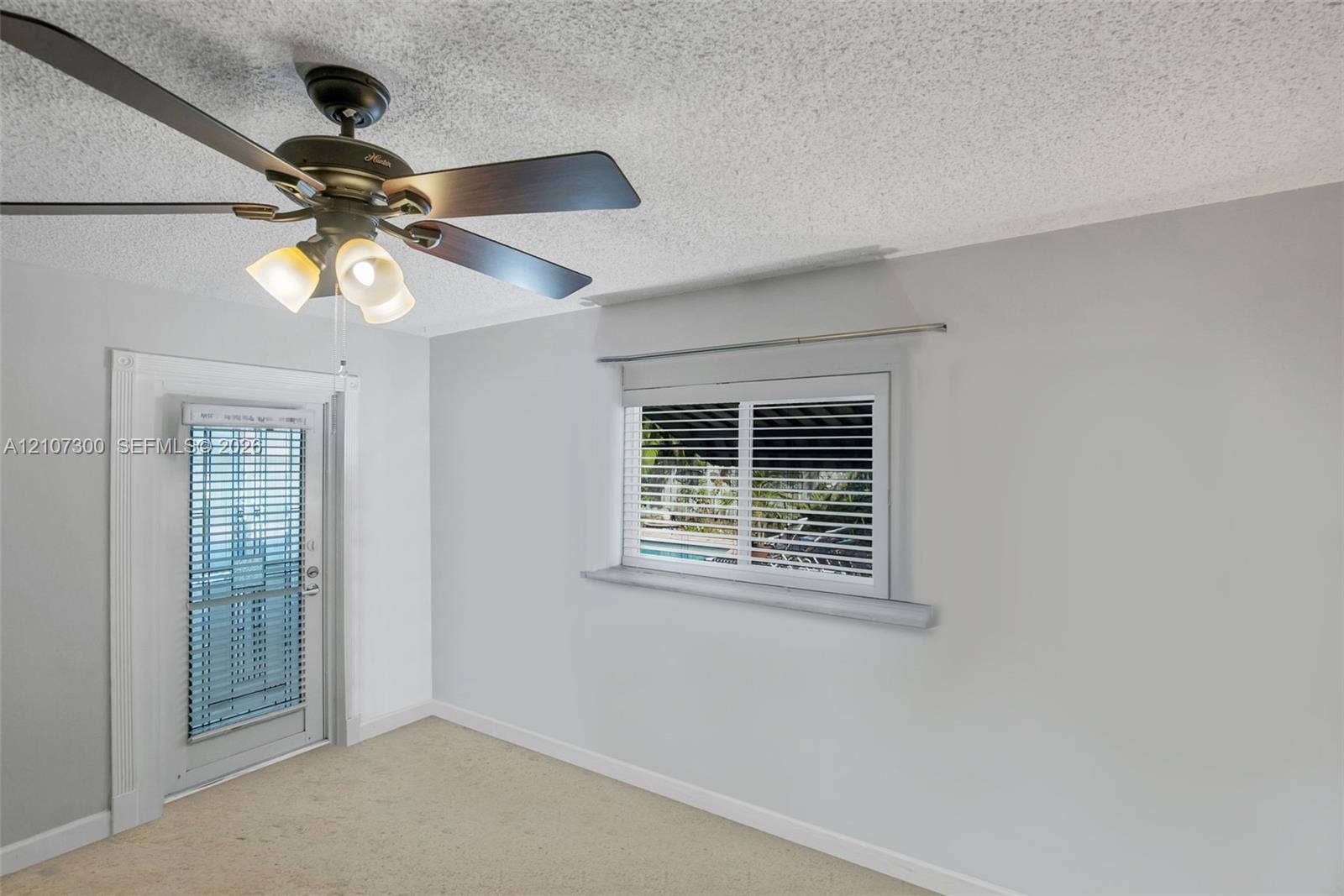 2051 NE 68th Street, Fort Lauderdale, FL 33308 Neutral interior featuring a ceiling fan with integrated three-light fixture, single-panel exterior door with glass insert, horizontal window blinds, and carpeted flooring