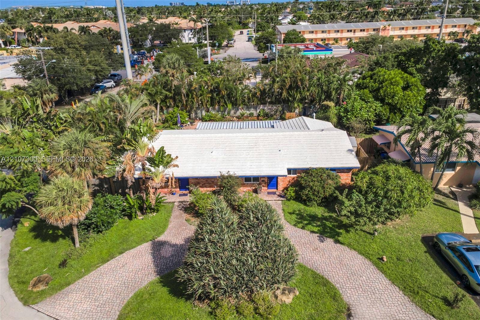 2051 NE 68th Street, Fort Lauderdale, FL 33308 Single-story residence featuring a brick exterior, white-tiled roof, and a circular paver driveway surrounded by mature landscaping