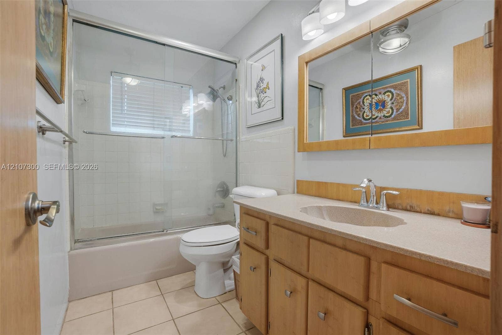 2051 NE 68th Street, Fort Lauderdale, FL 33308 Bathroom featuring wood cabinetry, a light-toned solid surface countertop with an integrated basin, a combination bathtub and shower with glass sliding doors, and a built-in medicine cabinet with integrated lighting