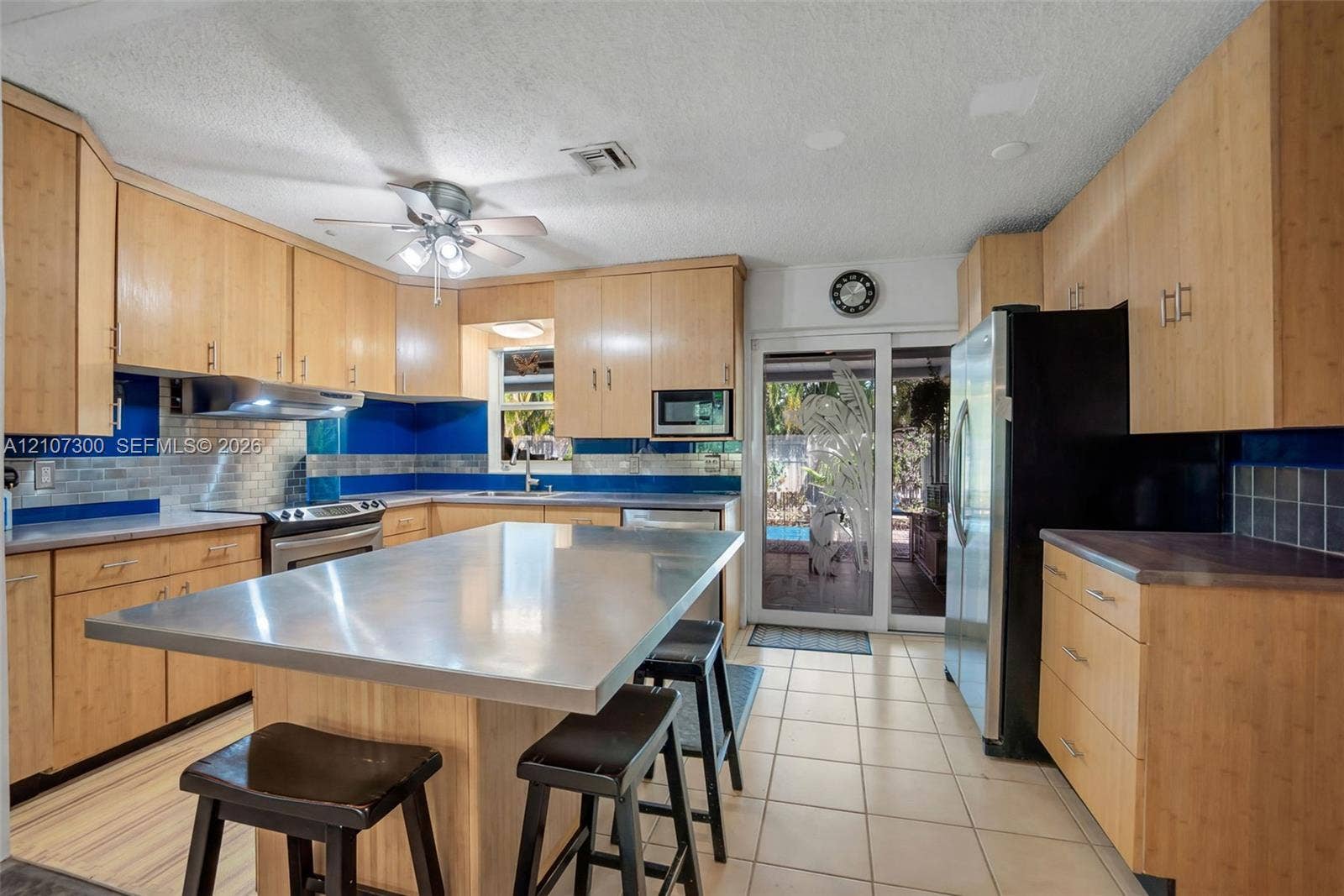 2051 NE 68th Street, Fort Lauderdale, FL 33308 Kitchen featuring light wood-finish cabinetry, a stainless steel island countertop, tile flooring, blue glass tile backsplash, and stainless steel appliances