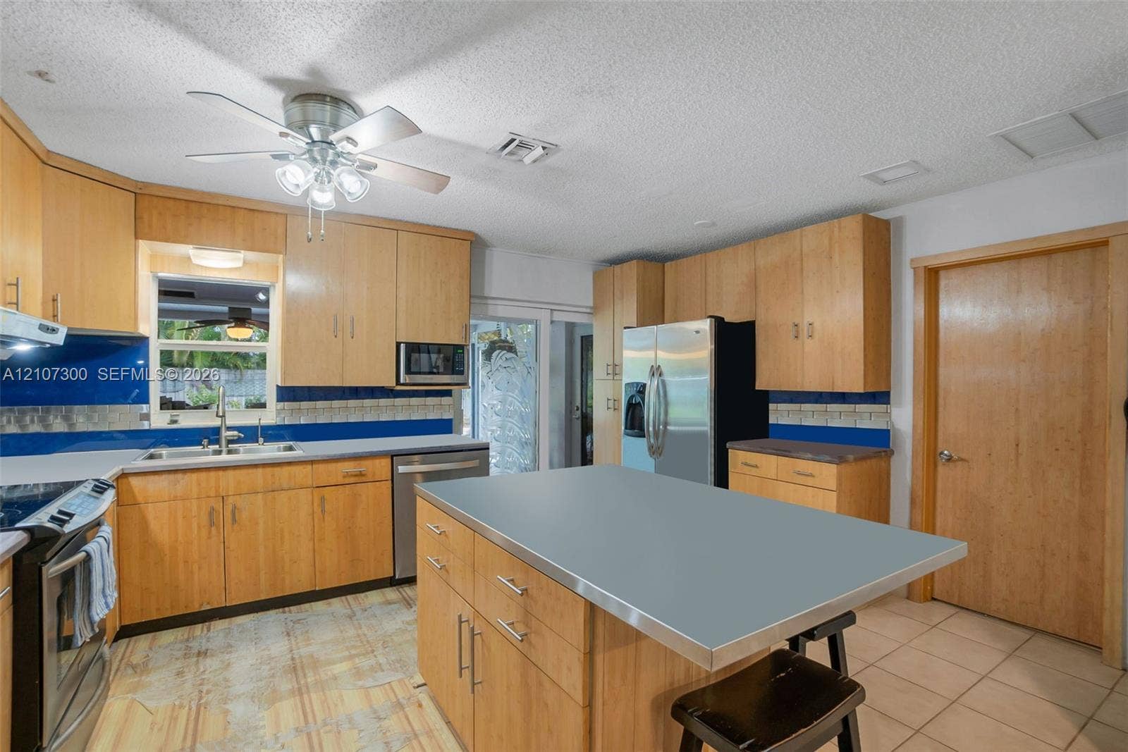 2051 NE 68th Street, Fort Lauderdale, FL 33308 Kitchen featuring light-finish cabinetry, a central island with a solid-surface countertop, stainless steel appliances, blue tiled backsplash, and tile flooring