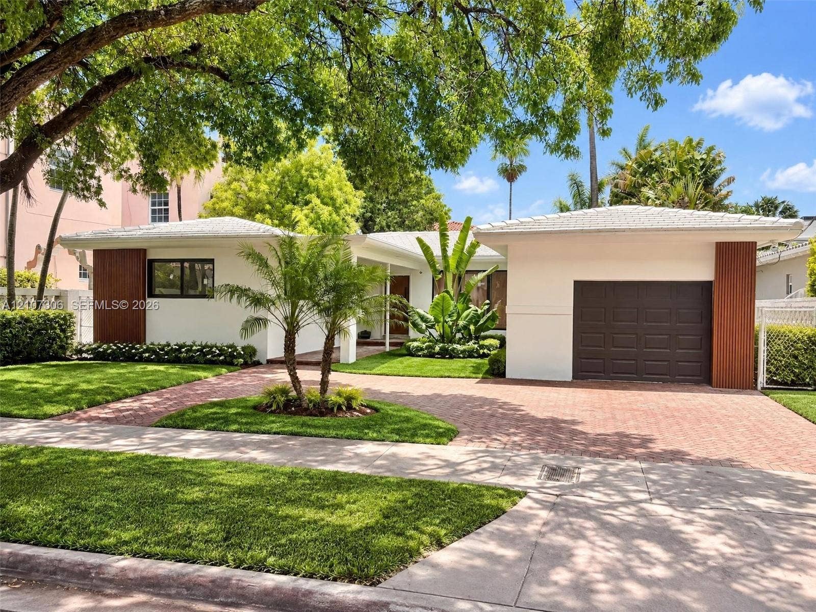 4322 Alton Road, Miami Beach, FL 33140 Single-story residence featuring a crisp white exterior with vertical wood-accented columns, a paved brick driveway, a dedicated garage, and mature landscaping