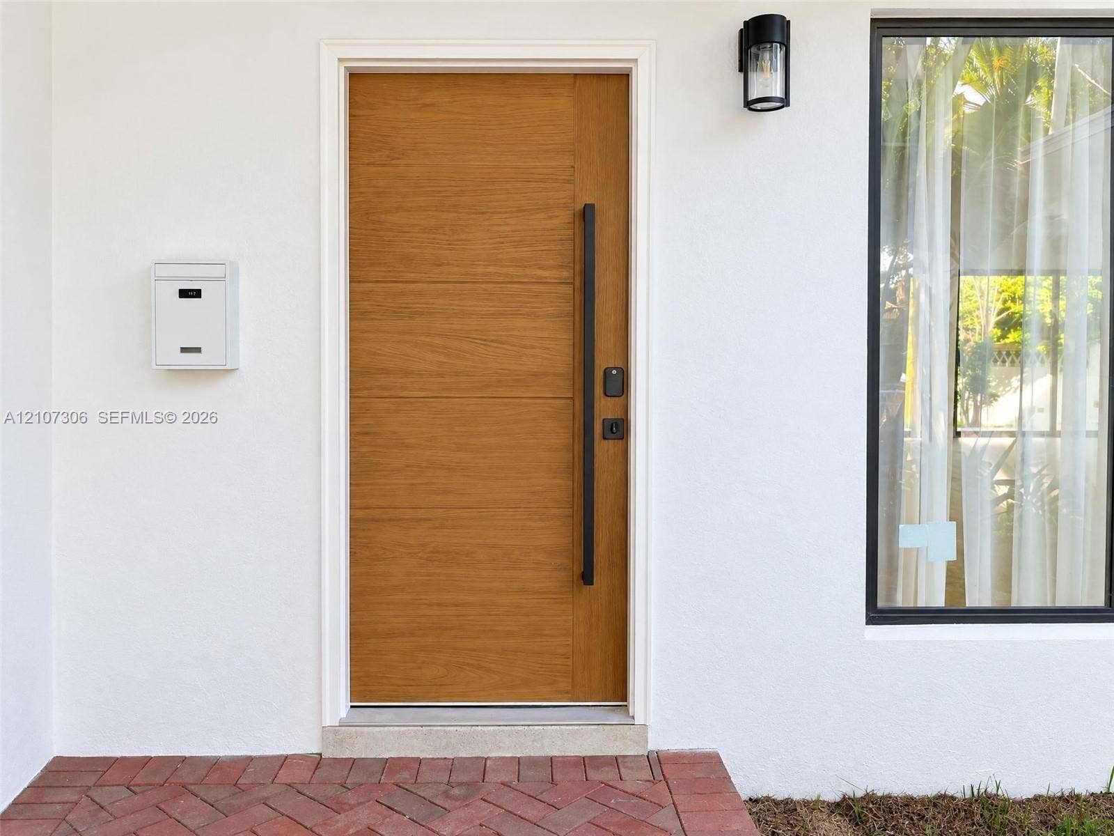 4322 Alton Road, Miami Beach, FL 33140 Wood-finish front door with a vertical matte black pull handle, white stucco exterior, exterior wall-mounted lighting, and herringbone-patterned brick entryway
