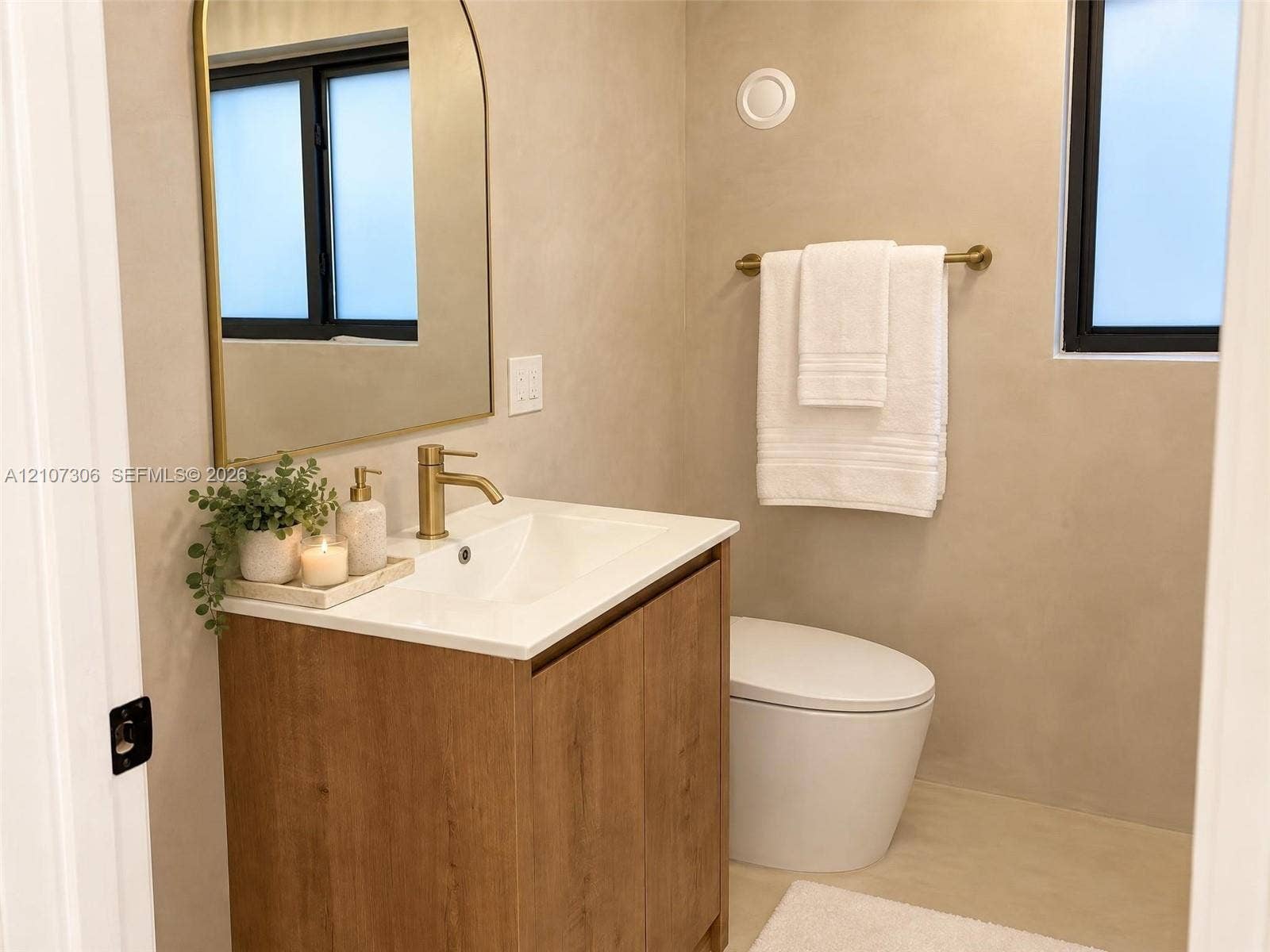 4322 Alton Road, Miami Beach, FL 33140 Wood-finish vanity cabinet with a white basin, arched wall mirror, brass-toned faucet, and hardware, complemented by textured wall finishes and a dual-flush toilet