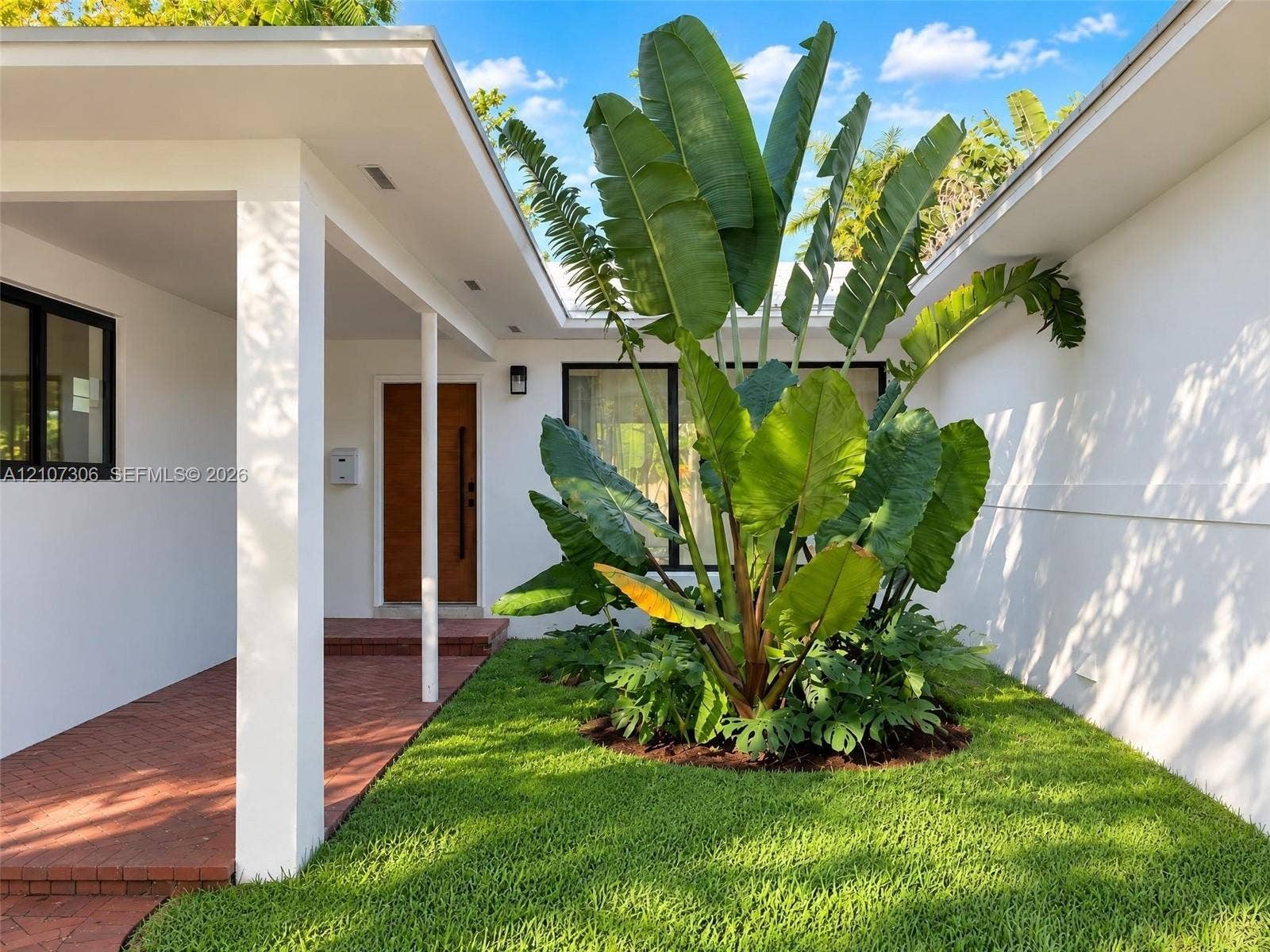 4322 Alton Road, Miami Beach, FL 33140 Courtyard entryway featuring a red brick floor, white stucco exterior walls, a natural wood-finish front door, and mature tropical landscaping