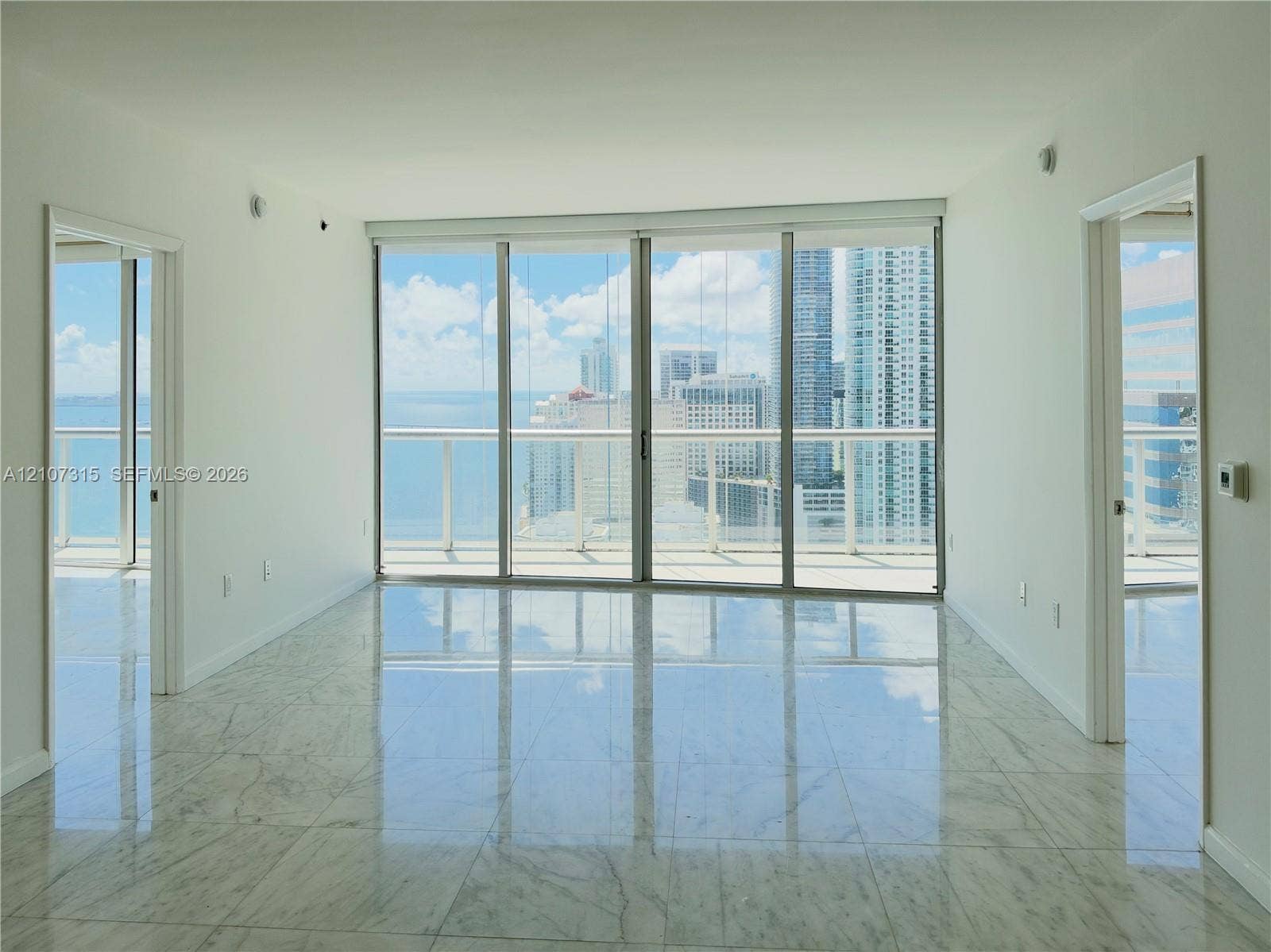 495 Brickell Avenue, #3803, Miami, FL 33131 Floor-to-ceiling glass sliding doors, stone-finish flooring, and an expansive balcony featuring a metal railing with glass panels, showcasing urban and water scenery