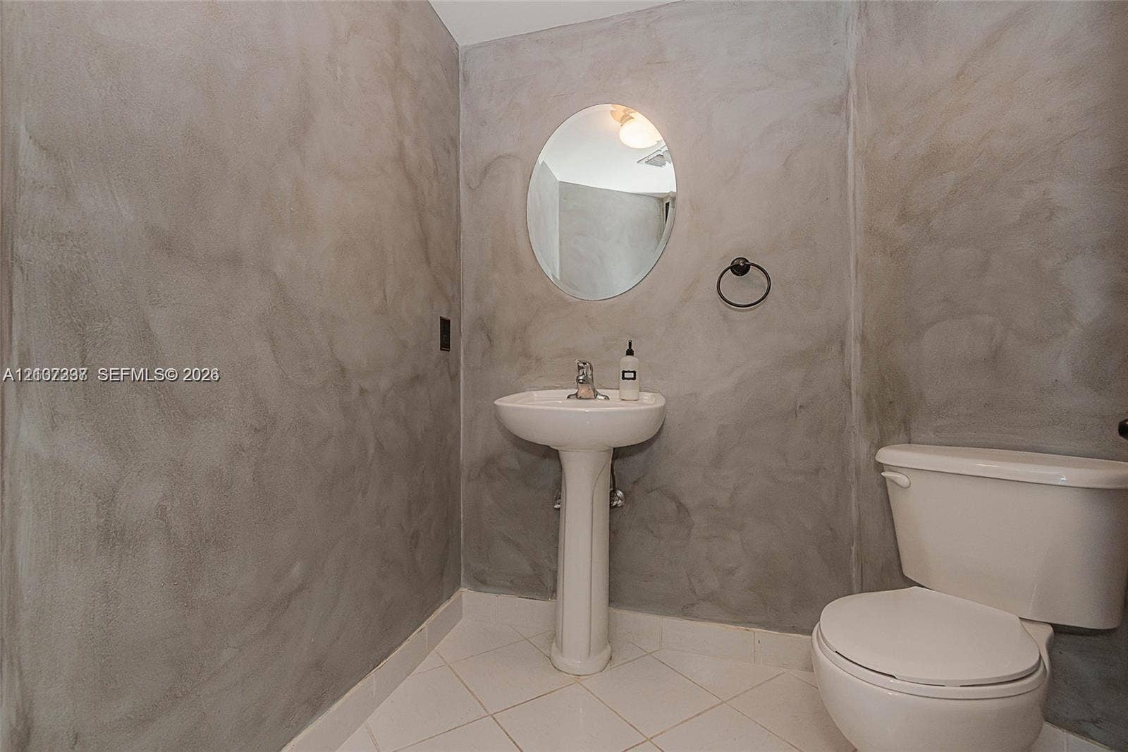 1881 79th St Causeway, #1007, North Bay Village, FL 33141 Textured concrete-finish walls, white ceramic pedestal sink, oval wall-mounted mirror, matte black towel ring, and ceramic tile flooring