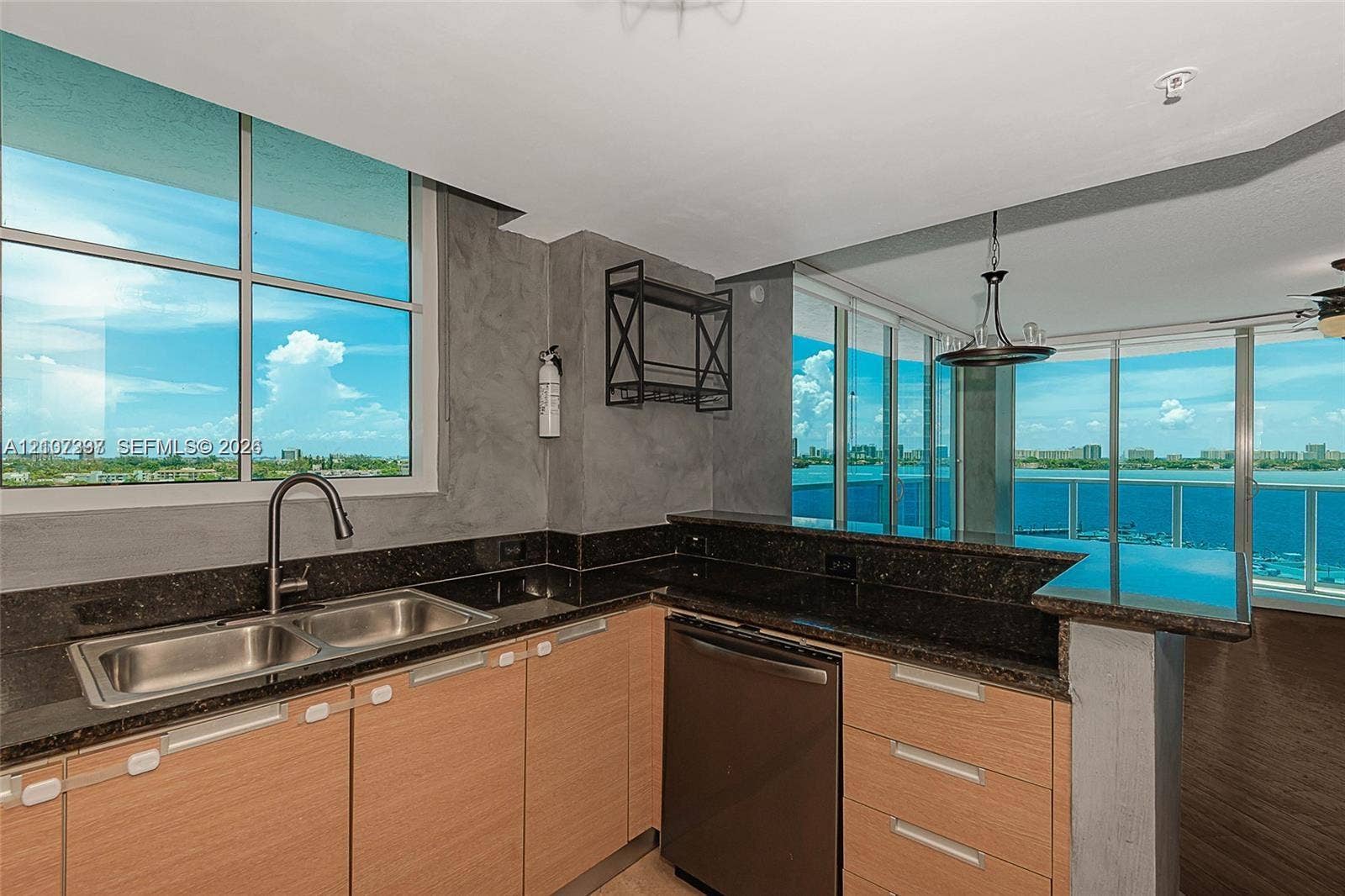 1881 79th St Causeway, #1007, North Bay Village, FL 33141 Kitchen featuring natural wood-finish cabinetry, dark stone countertops, stainless steel dual-basin sink, and built-in dishwasher, punctuated by expansive floor-to-ceiling windows