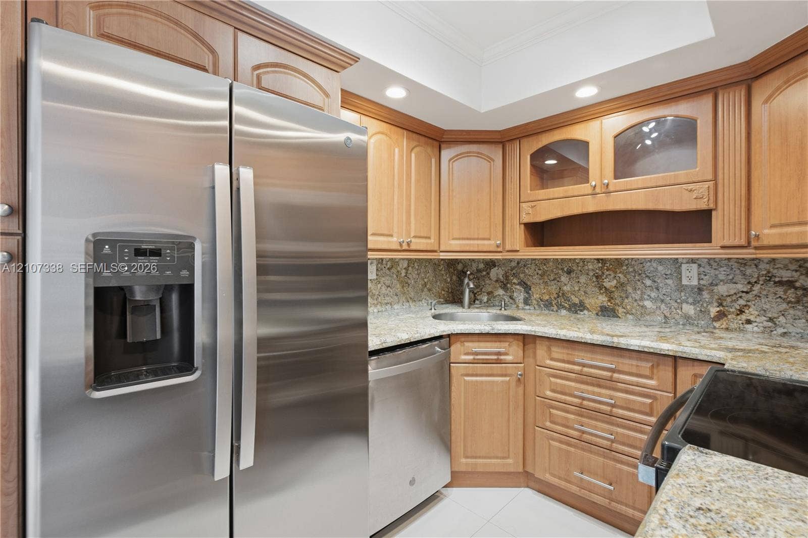 20355 NE 34th Court, #1824, Aventura, FL 33180 Stainless steel side-by-side refrigerator, natural stone countertops, tile backsplash, light wood-tone cabinetry with glass-front display accents, and recessed ceiling lighting