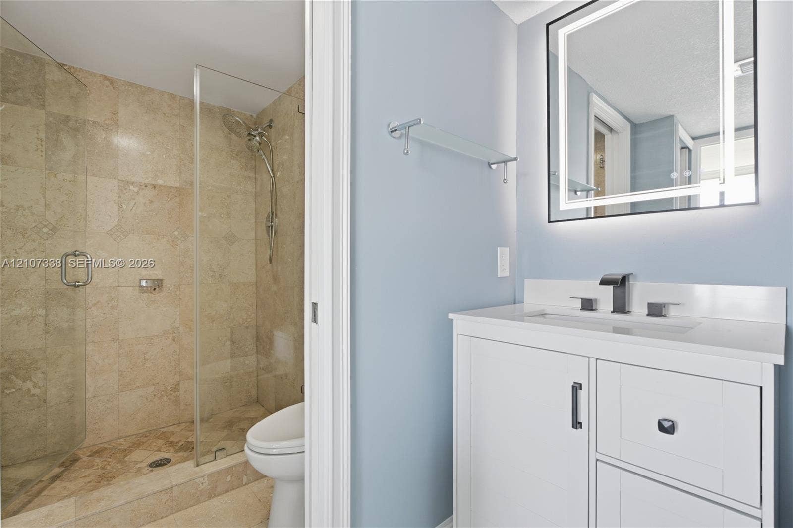 20355 NE 34th Court, #1824, Aventura, FL 33180 Bathroom featuring a white vanity with a quartz countertop, rectangular under-mount sink, matte black widespread faucet, and illuminated vanity mirror, alongside a walk-in shower with stone-tile surround and a glass door