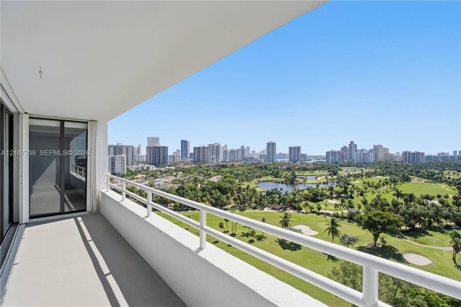 20355 NE 34th Court, #1824, Aventura, FL 33180 Covered balcony featuring white railings, neutral flooring, and glass sliding door access, showcasing expansive golf course and city skyline scenery