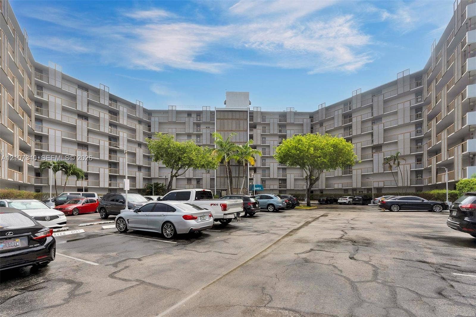 3401 N Country Club Drive, #406, Aventura, Aventura, FL 33180 Multi-story residential building featuring a U-shaped architectural design, balcony enclosures, surface parking, and mature palm trees