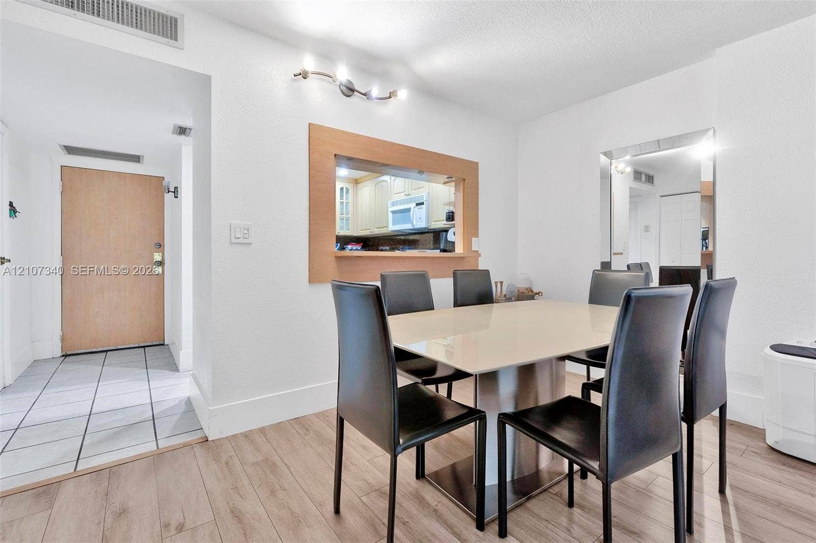 3401 N Country Club Drive, #406, Aventura, Aventura, FL 33180 Dining area featuring wood-finish flooring, a wall-mounted multi-bulb light fixture, a framed pass-through opening to the kitchen, and a floor-to-ceiling mirror