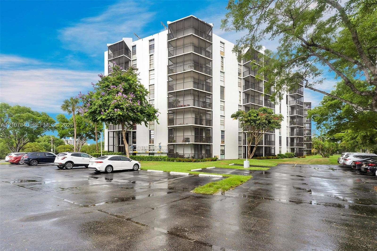 3401 N Country Club Drive, #406, Aventura, Aventura, FL 33180 Multi-story building exterior featuring white concrete facade, vertical rows of screened balconies, paved surface parking, and mature landscaping