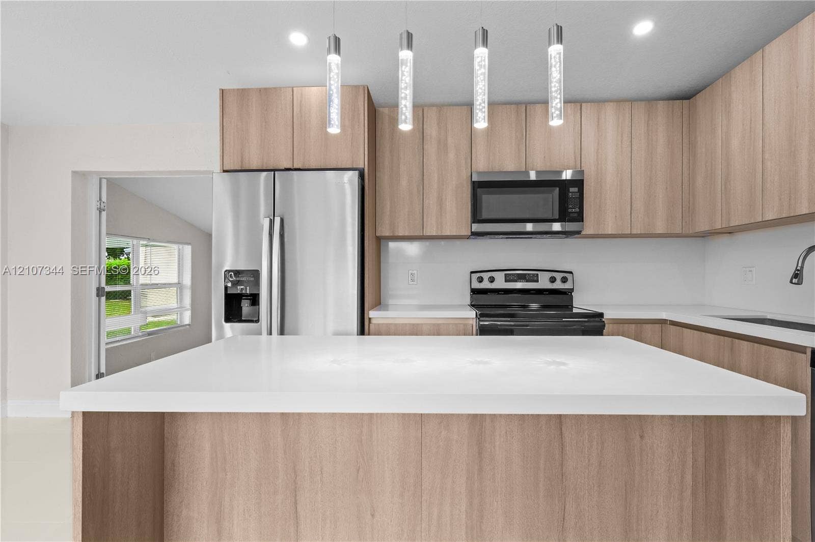 Kitchen featuring light wood-finish cabinetry, white solid-surface countertops, stainless steel appliances, and four cylindrical glass pendant lights